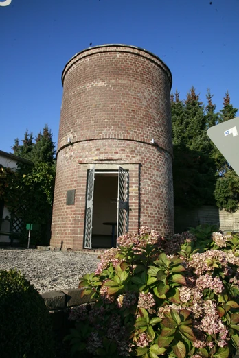 Neschen Wasserturm Alter Wasserturm aus Backstein, umgeben von Blumen und Bäumen, mit offenem Eingang und blauem Himmel.
