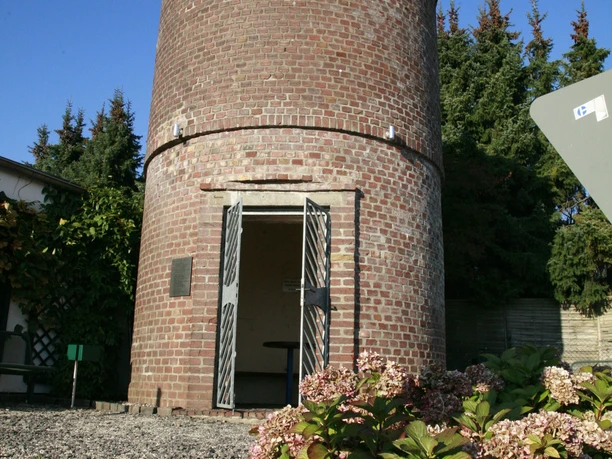 Neschen Wasserturm Alter Wasserturm aus Backstein, umgeben von Blumen und Bäumen, mit offenem Eingang und blauem Himmel.
