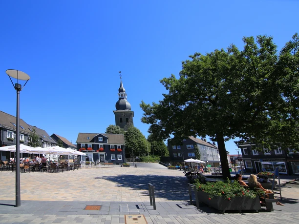 Marktplatz der Stadt Radevormwald Kirchplatz mit Bäumen, Tischen und Bar entfernt; Kirche mit Turm im Hintergrund, blauer Himmel.