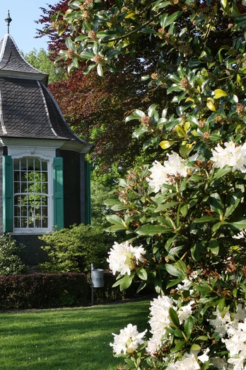 Kleines, historisches Gartenhaus mit grünem Fensterladen im Frühling, umgeben von blühenden Sträuchern.