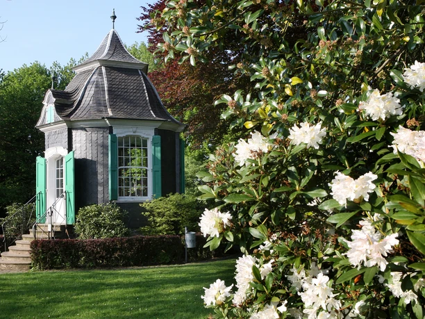 Rokkoko-Pavillon in Radevormwald Kleines, historisches Gartenhaus mit grünem Fensterladen im Frühling, umgeben von blühenden Sträuchern.