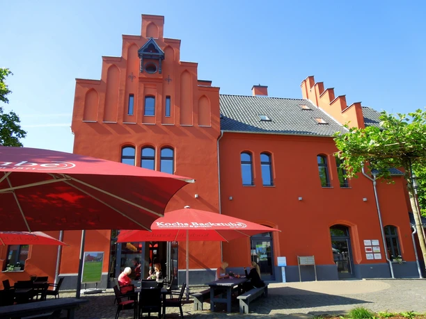 Wanderbahnhof Schladern (mit Touristinfo) Das Bild zeigt ein rotes, denkmalgeschütztes Gebäude mit Treppengiebeln, davor Außengastronomie.