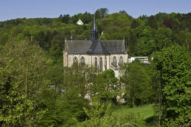 Altenberger Dom Gotische Kirche inmitten von üppigem Grün mit bewaldeten Hügeln im Hintergrund bei klarem Himmel.