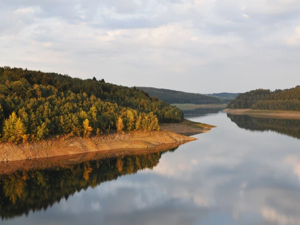 Blick auf die Dhünntalsperre Ein ruhiger, spiegelnder See umgeben von bewaldeten Hügeln unter einem leicht bewölkten Himmel.