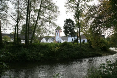 Die Dhünn in Odenthal Historisches Dorf mit Fachwerkhäusern und Kirchturm, eingebettet in üppige grüne Landschaft am Fluss.