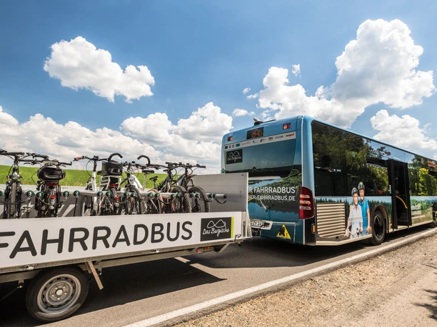Bergischer FahrradBus Ein moderner Bus mit Fahrradanhänger auf einer Landstraße unter blauem Himmel mit Wolken.