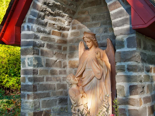 Heiligenhäuschen Steinskulptur eines Engels, der ein Kind schützt, unter einem kleinen, überdachten Steingewölbe.