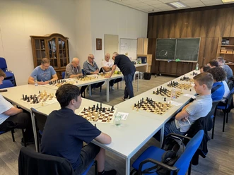 Simultanschachturnier mit Spielern an mehreren Schachbrettern auf weißen Tischen.