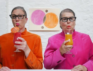 Kabarett Madamm Zwei Frauen mit Brille in bunter Kleidung stehen vor einer weißen Wand und trinken einen Cocktail mit einem Strohhalm.