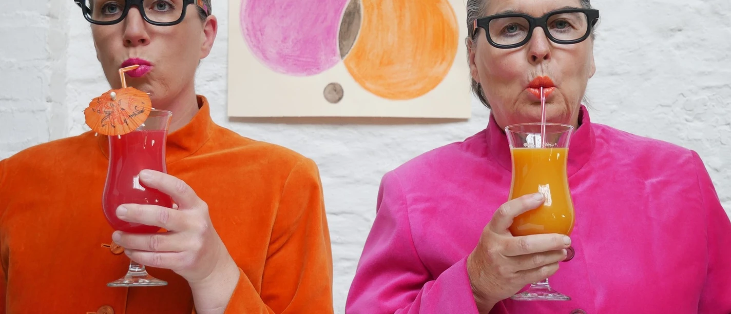 Kabarett Madamm Zwei Frauen mit Brille in bunter Kleidung stehen vor einer weißen Wand und trinken einen Cocktail mit einem Strohhalm.