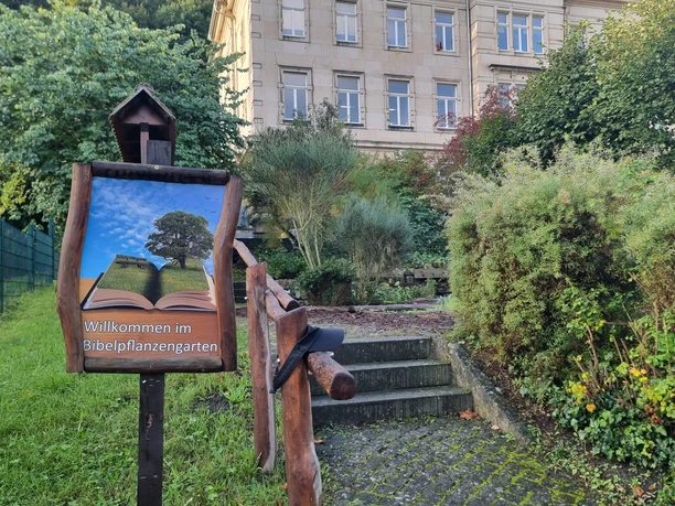 Bibelpflanzgarten Wegweiser zum Selbstpflückgarten mit einer offenen Buchillustration und einem Baum. Im Hintergrund ein Gebäude.