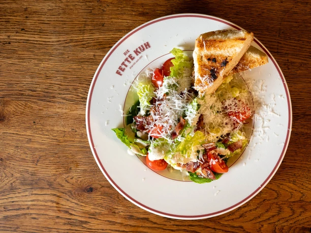 Die Fette Kuh - Caesar Salad Caesar salad with Parmesan, tomatoes, bacon, and toasted bread on a wooden table