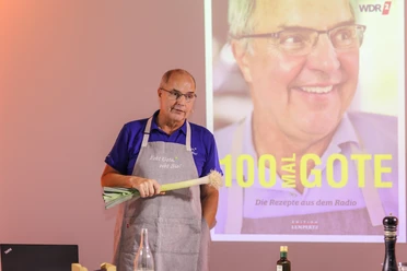 Helmut Gote Ein Mann steht mit einer Stange Lauch in der Hand auf einer Bühne. im Hintergrund läuft eine Präsentation.