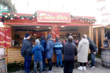 Dolce Vita | Märchenweihnachtsmarkt Kassel Vor dem Dolce Vita Stand stehen Menschen. Der Name der Hütte steht in gelber Schrift auf einem roten Schild.