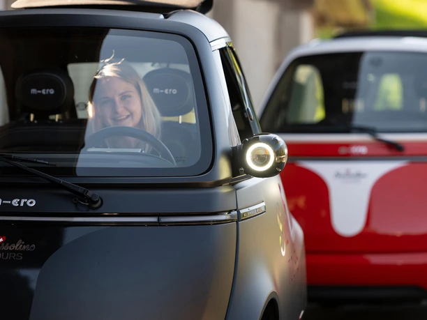 swissolino-fahrerin.jpg Lächelnde Fahrerin in schwarzem Swissolino-Microcar, rotes Fahrzeug dahinter