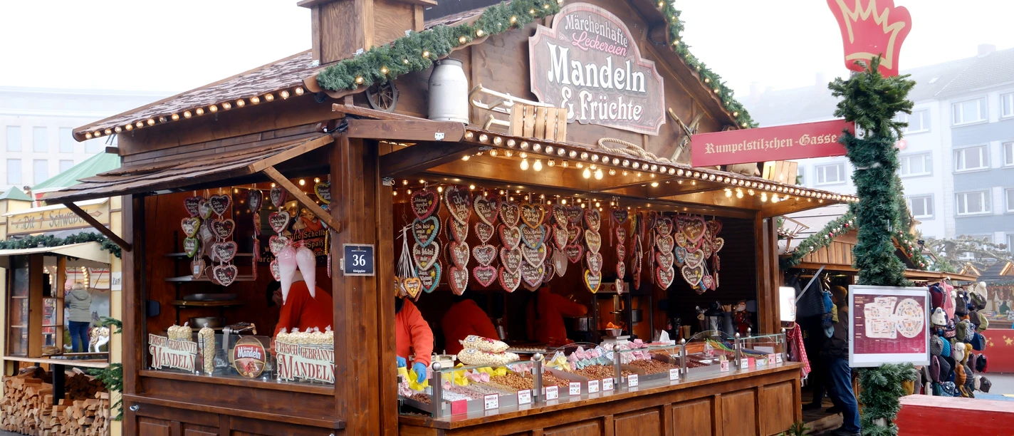 Mandeln & Früchte | Märchenweihnachtsmarkt Kassel Eine weihnachtlich geschmückte Holzhütte mit der Aufschrift "Mandeln & Früchte" ist dargestellt.