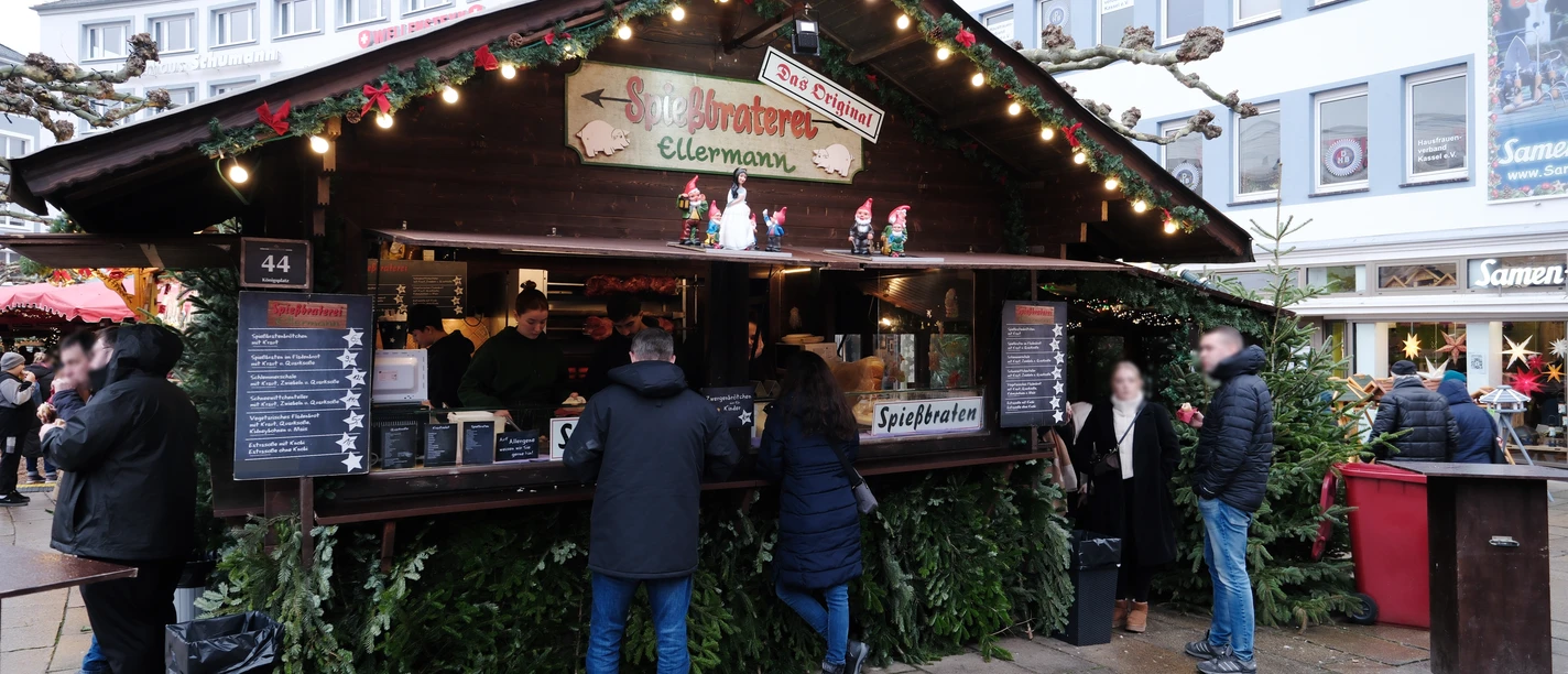 Vor der weihnachtlich verzierten Hütte mit dem Schild "Spießbraterei Ellermann" stehen einige Personen.