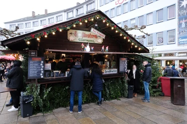 Speißbraten Ellermann | Märchenweihnachtsmarkt Kassel Vor der weihnachtlich verzierten Hütte mit dem Schild "Spießbraterei Ellermann" stehen einige Personen.