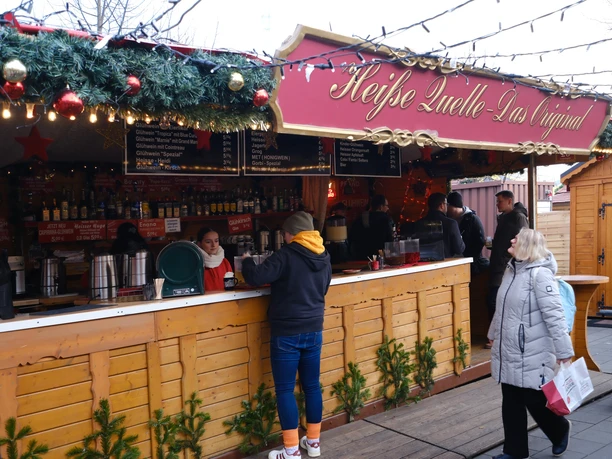 Stand Heiße Quelle | Märchenweihnachtsmarkt Kassel Die hölzerne Weihnachtsmarkthütte hat ein Schild mit dem Standnamen "Heiße Quelle".