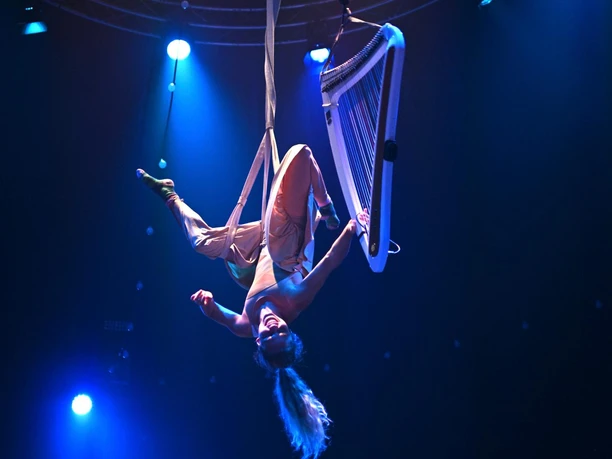 Cirque Bouffon Harmonia - The Christmas Show An aerial artist hangs upside down from silks next to a harp in the blue stage lighting