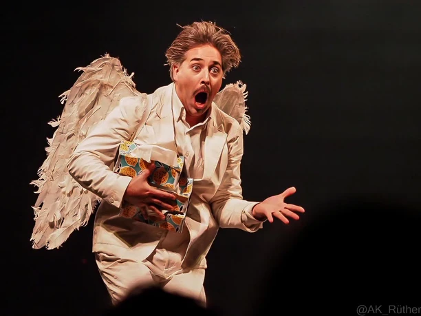 Cirque Bouffon Harmonia - The Christmas Show An artist in a white angel costume holds a gift on a dark circus stage.