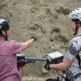 Saurierfährten in Bad Essen Zwei Radfahrer betrachten interessiert Fossilienspuren in einer Felswand.