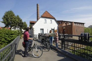 Radwanderer vor dem Tuchmacher Museum in Bramsche Drei Radfahrer studieren eine Karte vor einer historischen Wassermühle an einem sonnigen Tag.