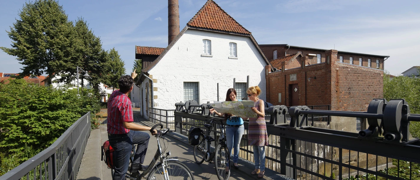 Radwanderer vor dem Tuchmacher Museum in Bramsche Drei Radfahrer studieren eine Karte vor einer historischen Wassermühle an einem sonnigen Tag.