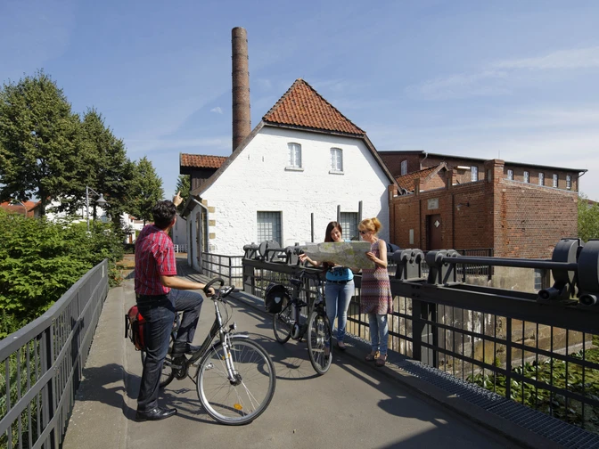Radwanderer vor dem Tuchmacher Museum in Bramsche Drei Radfahrer studieren eine Karte vor einer historischen Wassermühle an einem sonnigen Tag.