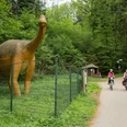 Die Sauerierfährten in Bad Essen Radfahrer auf einem Waldweg passieren eine Dinosaurier-Statue hinter einem Zaun.