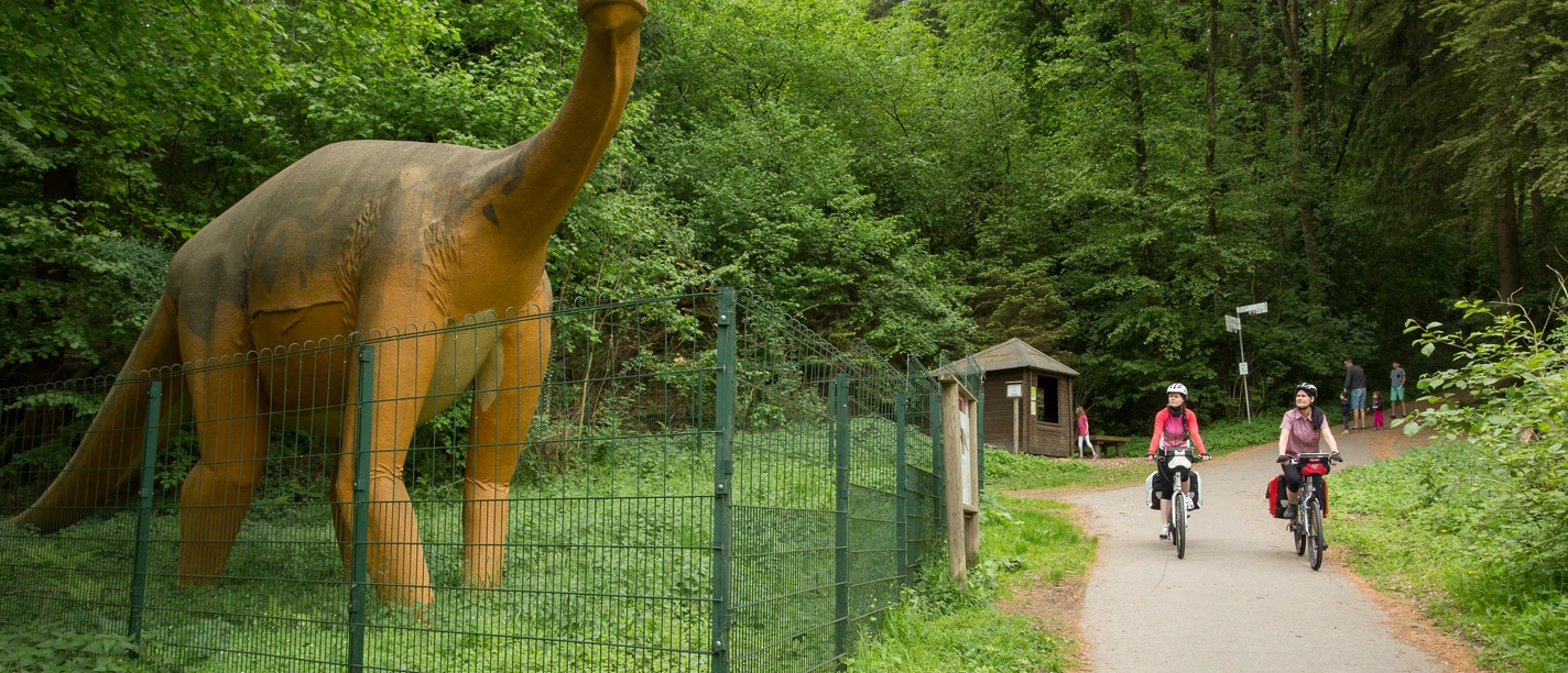 Die Sauerierfährten in Bad Essen Radfahrer auf einem Waldweg passieren eine Dinosaurier-Statue hinter einem Zaun.