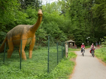 Die Sauerierfährten in Bad Essen Radfahrer auf einem Waldweg passieren eine Dinosaurier-Statue hinter einem Zaun.