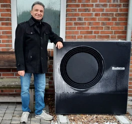Andreas Stampa neben Wärmepumpe Andreas Stampa steht vor einer Backsteinwand neben einer schwarzen Wärmepumpe.