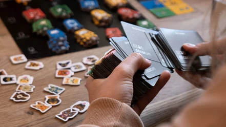 Speedgaming.jpg Eine Person schaut Spielkarten in ihrer Hand an. Auf dem Tisch im Hintergrund ist ein Spielbrett aufgebaut.