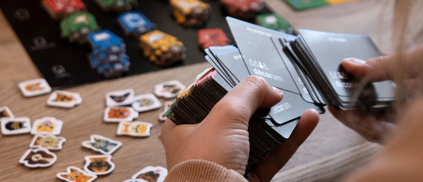 Speedgaming.jpg Eine Person schaut Spielkarten in ihrer Hand an. Auf dem Tisch im Hintergrund ist ein Spielbrett aufgebaut.