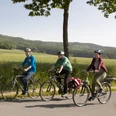 Unterwegs im Osnabrücker Land Drei Radfahrer fahren auf einer Straße durch eine weite grüne Landschaft bei sonnigem Wetter.