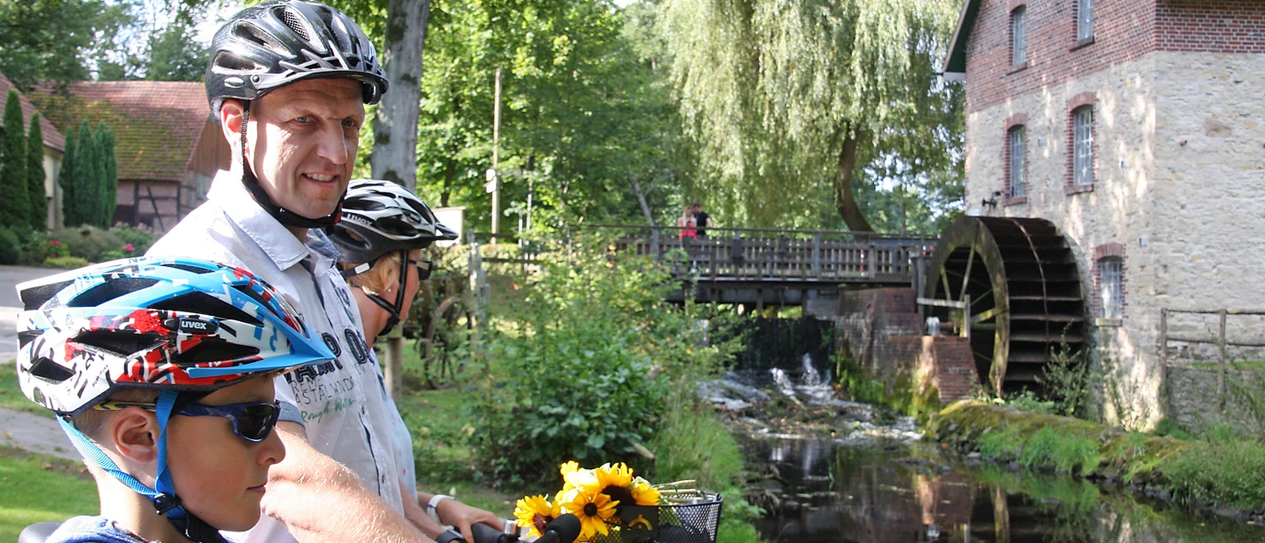 Radfahrer an der Nackten Mühle Eine Familie auf Fahrrädern vor einer historischen Wassermühle und einem ruhigen Bach.