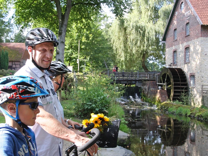 Radfahrer an der Nackten Mühle Eine Familie auf Fahrrädern vor einer historischen Wassermühle und einem ruhigen Bach.