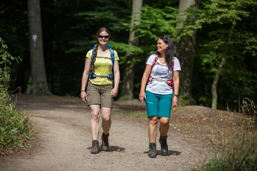 Twee wandelende vrouwen op een zonnig bospad, lachend, met rugzakken.