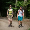 Wandern im Natur- und Geopark TERRA.vita Zwei wandernde Frauen in einem sonnigen Waldweg, lächelnd, mit Rucksäcken.