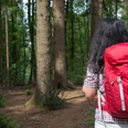 Vrouw met rode rugzak loopt over een bospad tussen hoge bomen.