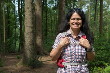 Vrouw met rugzak glimlacht in het zomerse bos voor hoge bomen en groen gebladerte.