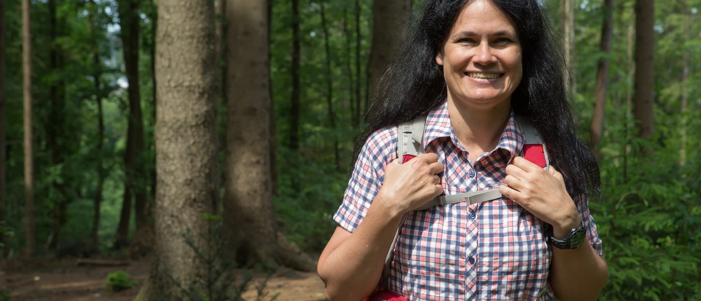 Unterwegs im Wald Frau mit Rucksack lächelt im sommerlichen Wald vor hohen Bäumen und grünem Laub.