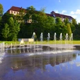 Springbrunnen vor einem weitläufigen, historischen Gebäude mit rotem Dach und grüner Umgebung.