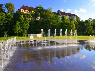 Kneipp-Erlebnispark in Bad Iburg: die Lehre von Sebastian Kneipp neu interpretiert. Springbrunnen vor einem weitläufigen, historischen Gebäude mit rotem Dach und grüner Umgebung.