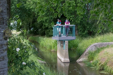 Twee wandelaars staan op een uitkijkplatform boven een riviertje op het platteland.