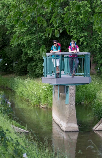 Bifurkation Zwei Wanderer stehen auf einer Aussichtsplattform über einem kleinen Fluss im Grünen.