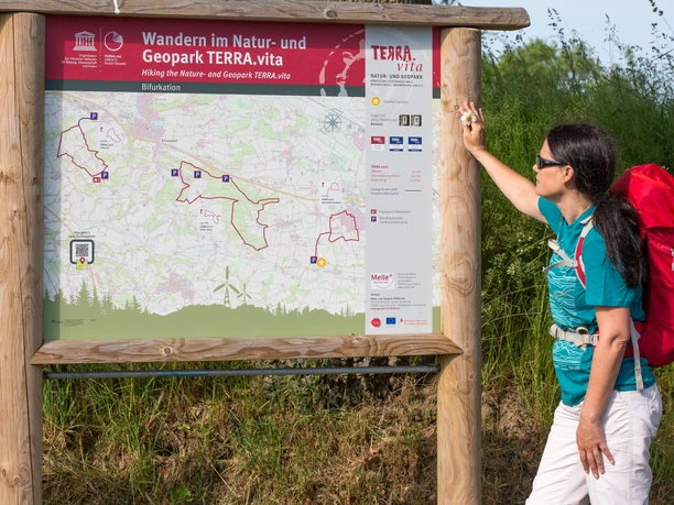 Infotafel En vandrer studerer et kort over TERRA.vita Geopark på et træskilt i landskabet.