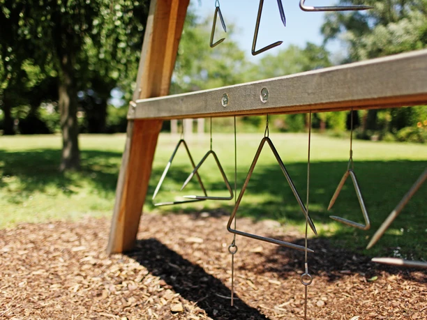 klang-und-rhythmusgarten-ein-design Metall-Dreiecke hängen an einem hölzernen Gestell im sonnigen Freien auf einer Grünfläche.