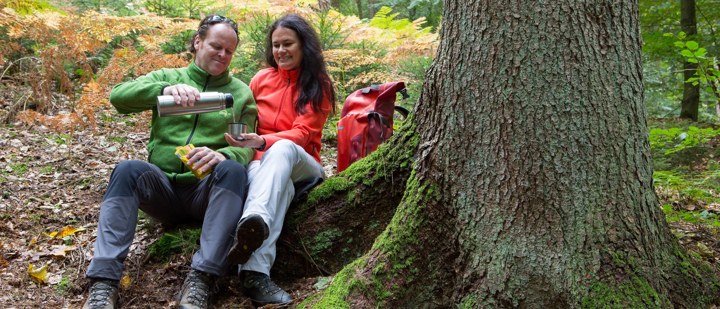 Een stel in outdoorkleding geniet van een pauze in het bos en schenkt thee in een mok.